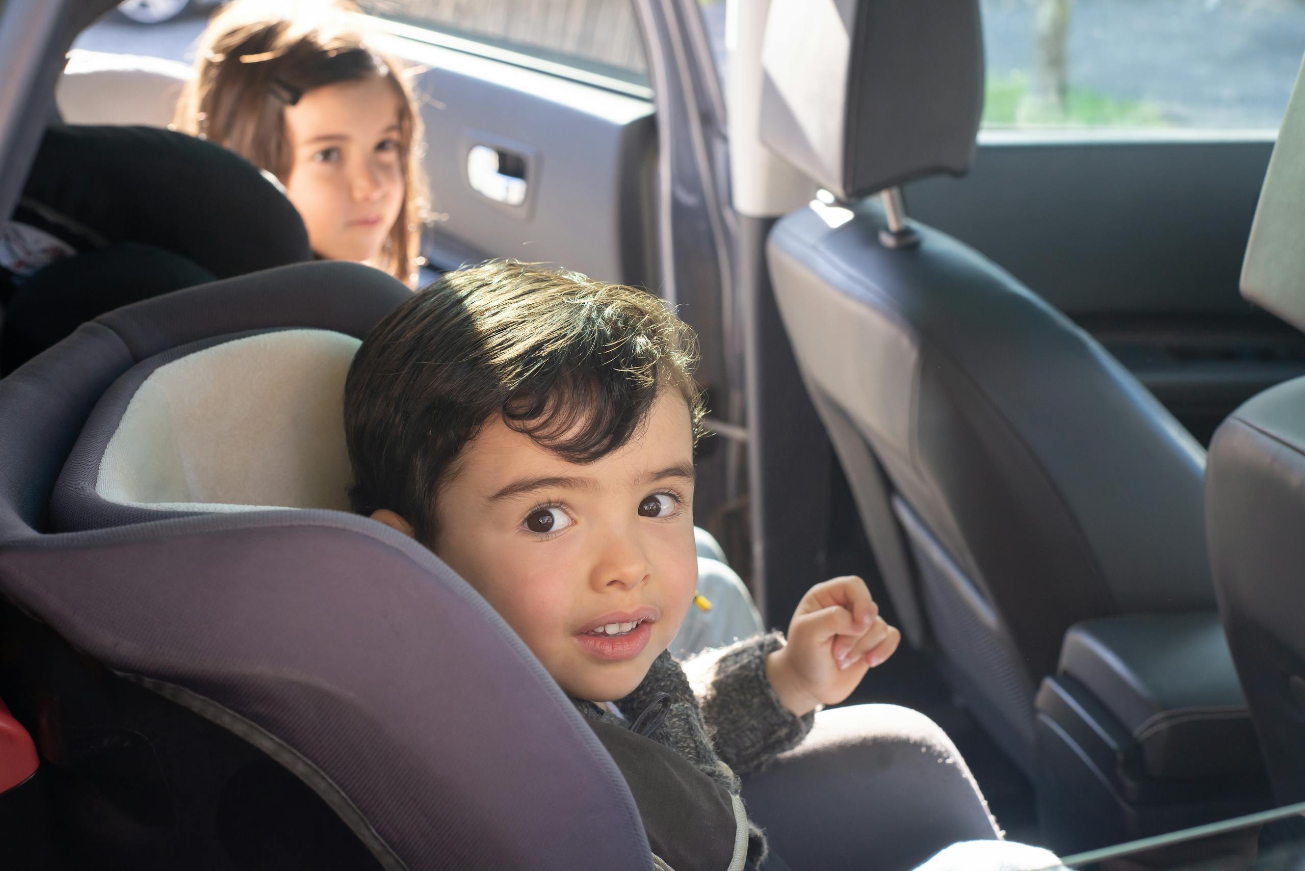 A toddler sitting in a car seat while another child is getting into the car.